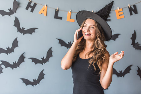 Beautiful young woman in witch hat is talking on the mobile phone and smiling, on background...