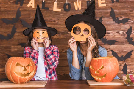 Cute little girl and her beautiful mom in witch hats are looking at camera and smiling while sitting on wooden background decorated for Halloweenの写真素材