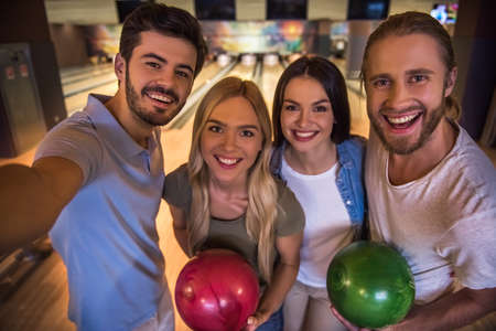 Happy young friends are holding balls, doing selfie and smiling while playing bowling togetherの写真素材