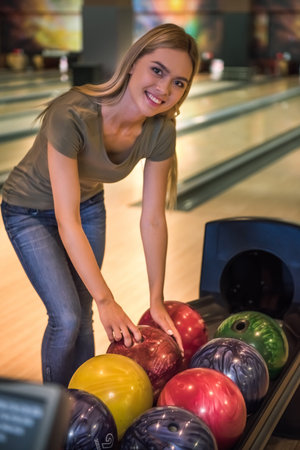 Attractive blonde girl is choosing a bowling ball, looking at camera and smiling ready to play bowlingの写真素材