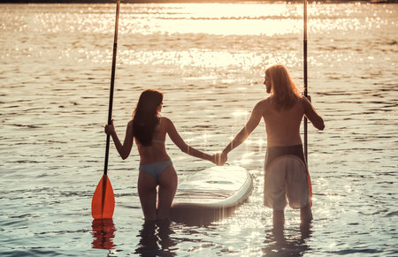 Back view of beautiful young couple holding hands and paddles, going to stand up paddle boardの写真素材