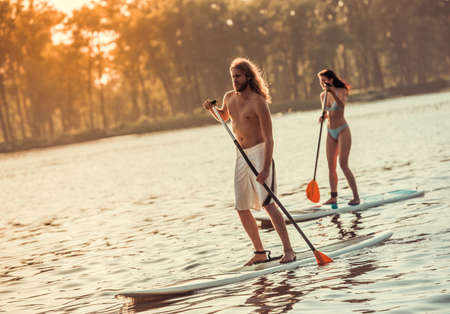Beautiful young couple is SUP surfing on the river, sun is going downの写真素材