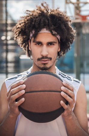 Attractive mulatto basketball player is holding a ball and looking at camera while standing on basketball court outdoorsの写真素材