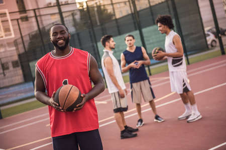 Handsome basketball players are talking and smiling on basketball court outdoors, Afro American guy in the foreground is looking at cameraの写真素材
