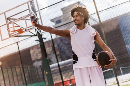 Attractive mulatto basketball player is doing selfie using a smart phone and smiling while standing on basketball court outdoorsの写真素材