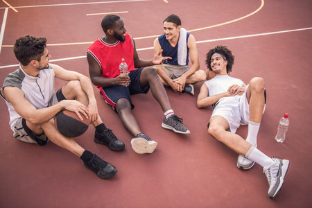 Handsome basketball players are talking and smiling while sitting on basketball court outdoors after the gameの写真素材