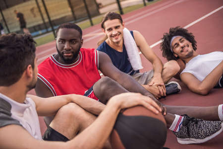 Handsome basketball players are talking and smiling while sitting on basketball court outdoors after the gameの写真素材