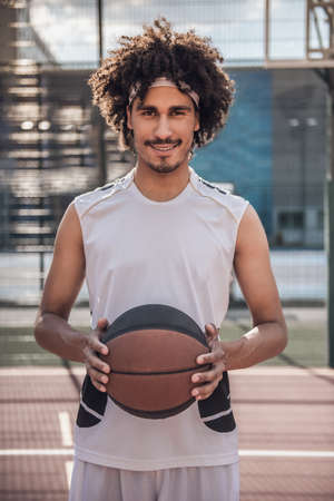 Attractive mulatto basketball player is holding a ball, looking at camera and smiling while standing on basketball court outdoorsの写真素材