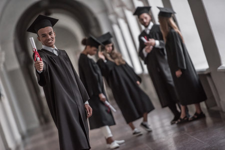Successful graduates in academic dresses are talking in university hall, guy in the foreground is...