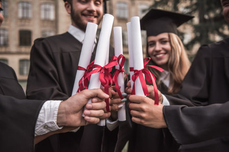 Cropped image of successful graduates in academic dresses holding diplomas and smiling while standing outdoorsの写真素材