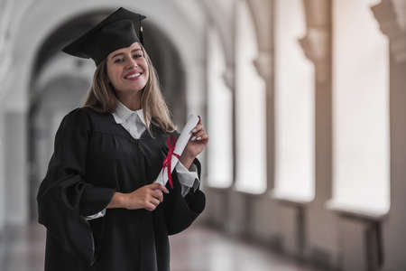 Attractive young female graduate in academic dress is holding a diploma, looking at camera and smiling while standing in university hallの写真素材