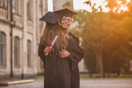 Beautiful female graduates in academic dresses are hugging and smiling while standing outdoorsの写真素材