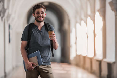 Attractive young student is holding a cup of coffee, looking at camera and smiling while standing in university hallの写真素材