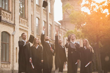 Successful graduates in academic dresses are holding diplomas, throwing their caps and smiling while standing outdoorsの写真素材