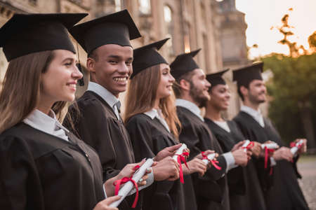 Successful graduates in academic dresses are holding diplomas, looking forward and smiling while standing in a row outdoorsの写真素材