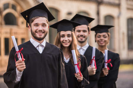 Successful graduates in academic dresses are holding diplomas, looking at camera and smiling while standing outdoorsの写真素材