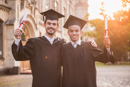 Handsome graduates in academic dresses are holding diplomas, looking at camera and smiling while standing outdoorsの写真素材