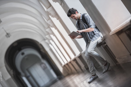 Handsome young student is using a digital tablet and smiling while sitting on the window-sill in university hallの写真素材