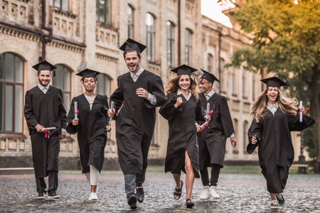 Successful graduates in academic dresses are holding diplomas, looking at camera and smiling while running outdoorsの写真素材