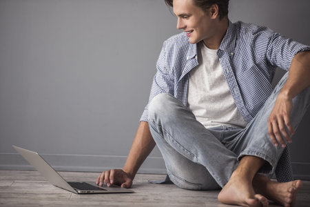 Handsome young man in casual clothes is using a laptop and smiling while sitting on the floor at homeの写真素材