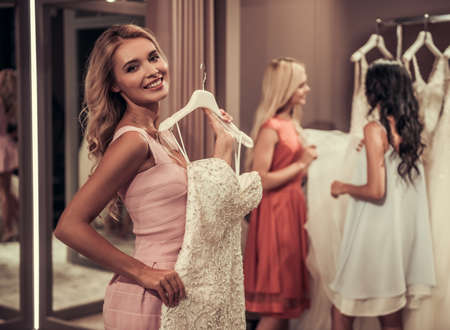 Beautiful young women are choosing elegant wedding dresses in modern wedding salon, blonde in the foreground is looking at camera and smilingの写真素材