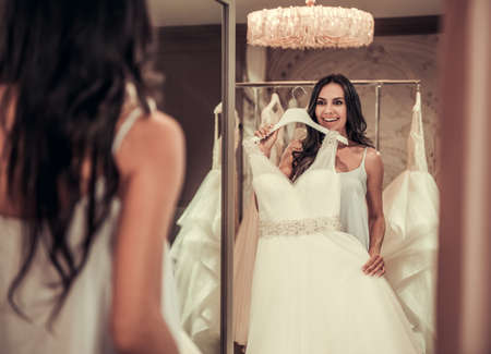 Beautiful young bride is looking into the mirror and smiling while choosing elegant wedding dresses in modern wedding salonの写真素材
