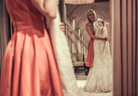 Attractive young bride is looking into the mirror and smiling while choosing elegant wedding dresses in modern wedding salonの写真素材
