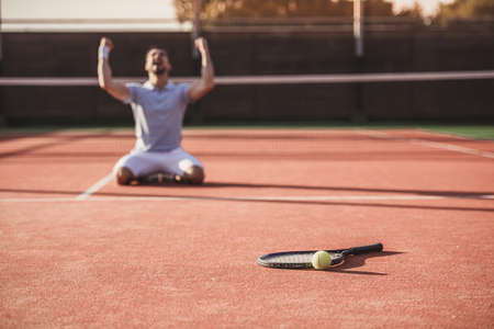 Handsome man is standing on his knees and screaming with happiness while playing tennis on court outdoorsの写真素材