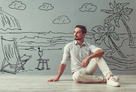 Handsome man in smart casual clothes is looking away, dreaming and smiling while sitting on the floor next to the wall with drawn seasideの写真素材