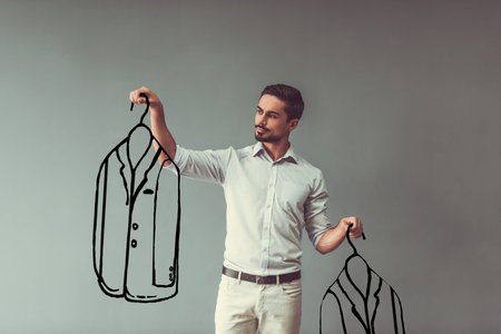 Handsome man in smart casual wear is holding drawn men suits and choosing between, on gray backgroundの写真素材