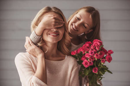 Pretty teenage daughter is holding flowers and covering her mom's eyes while making a surprise, both are smilingの写真素材
