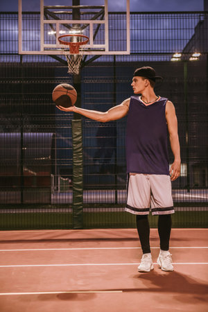Full length portrait of stylish young basketball player in cap standing with a ball on basketball court outdoors in the eveningの写真素材