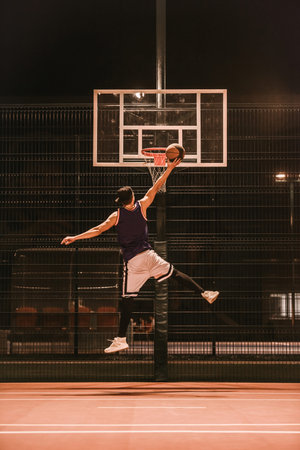Full length portrait of stylish young basketball player in cap jumping and shooting a ball through the hoop while playing outdoors at nightの写真素材
