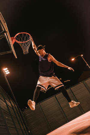 Full length portrait of stylish young basketball player in cap jumping and shooting a ball through the hoop while playing outdoors at nightの写真素材
