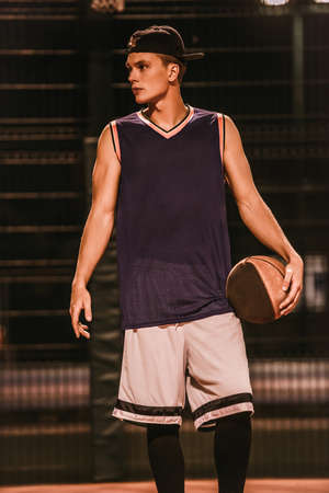 Stylish young basketball player in cap is standing with a ball on basketball court outdoors at nightの写真素材
