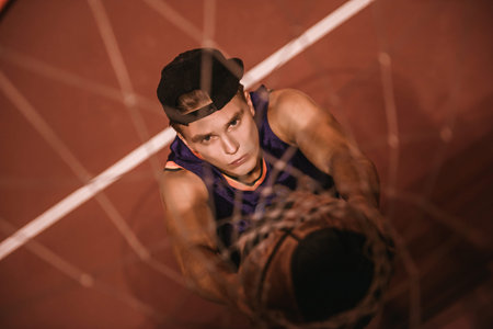 Top view of stylish young basketball player in cap shooting a ball through the hoop while playing outdoors at nightの写真素材