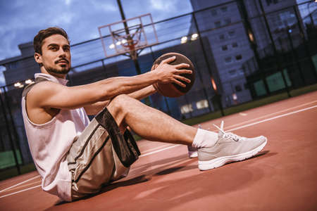 Handsome basketball player is sitting with a ball on basketball court outdoor in the eveningの写真素材