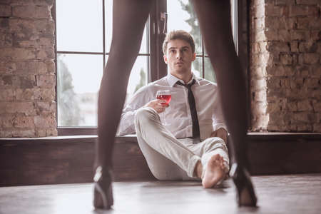 Handsome young businessman in suit is holding a glass of wine and looking at the girl while sitting on the floor, girl's legs in the foregroundの写真素材