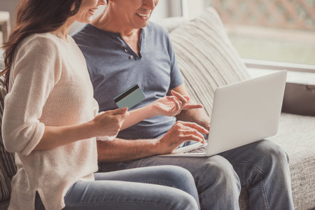 Beautiful couple is doing shopping online using a laptop and smiling while sitting on couch at home,...