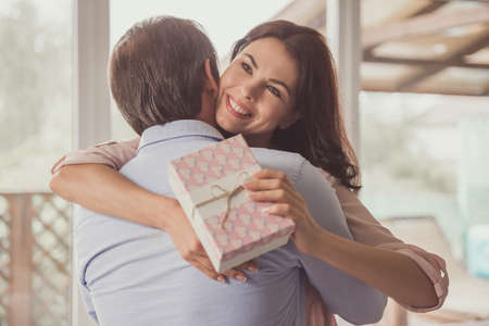 Couple at home. Handsome mature man is giving a present to his beautiful wife, she is hugging him and smilingの写真素材