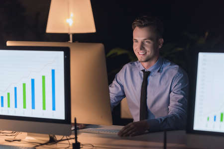 Handsome businessman in classic suit is using a computer and smiling while working in office late in the eveningの写真素材