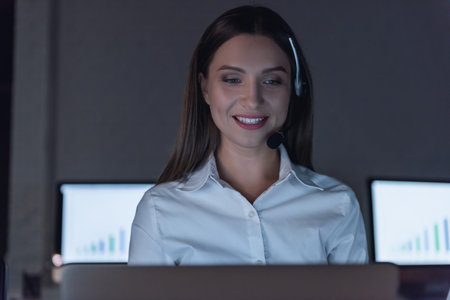 Beautiful business woman in headset is using a computer and smiling while working in office late at...