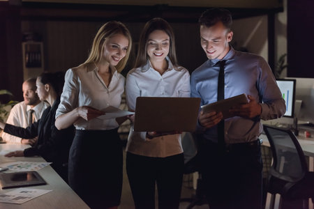 Successful business people are using gadgets and smiling while working in office late at night, colleagues in the backgroundの写真素材