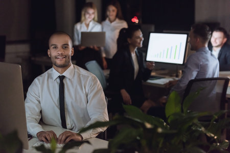Handsome Afro American businessman is looking at camera and smiling while working with a computer in office late at night, his colleagues nearの写真素材