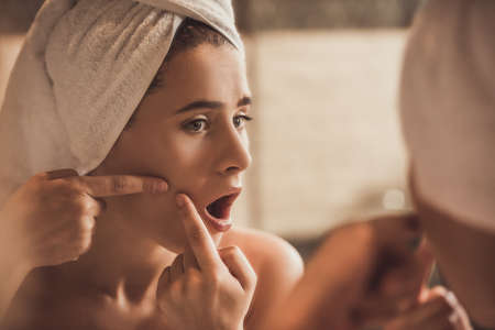 Young woman with towel on her head is squeezing pimples on her cheek while looking into the mirror in bathroomの写真素材