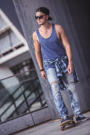 Stylish guy in casual clothes, cap and glasses is going skateboarding in the cityの写真素材