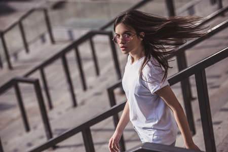 Stylish dark-haired girl in casual clothes and glasses is looking at camera while going down the stairs outdoorsの写真素材