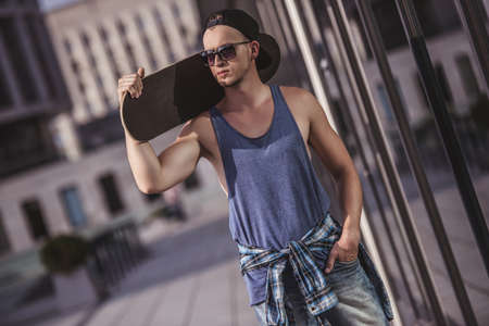 Stylish guy in casual clothes, cap and glasses is holding a skateboard and looking away while walking in the cityの写真素材