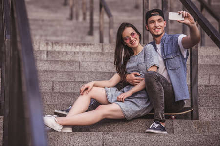 Stylish young couple is doing selfie using a phone and smiling while sitting on the skateboard on stairsの写真素材