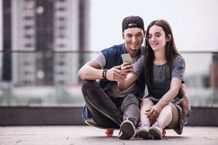 Stylish young couple is hugging, using a smart phone and smiling while sitting on the skateboard outdoorsの写真素材
